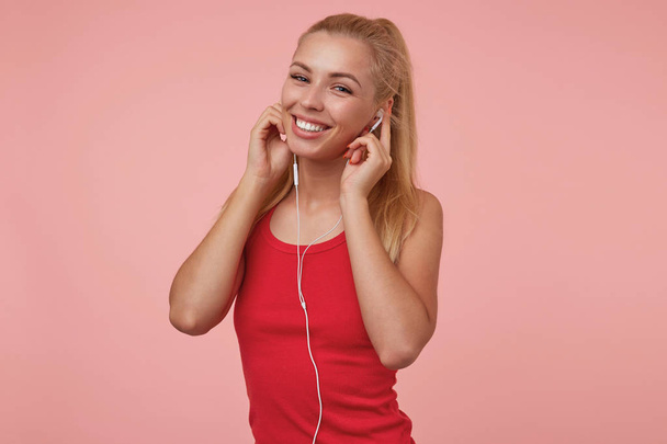 Indoor shot de mujer rubia feliz en ropa casual insertando auriculares en los oídos, sonriendo sinceramente y mirando a la cámara, de pie sobre el fondo rosa
 - Foto, imagen