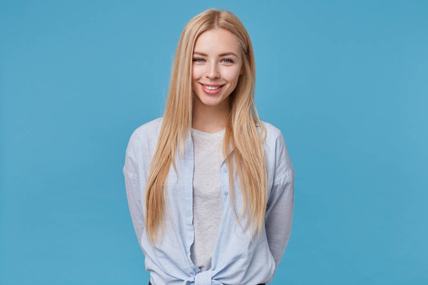 Positive attractive young blonde lady in casual clothes isolated over blue background, looking at camera cheerfully and showing her pleasant emotions - Фото, изображение