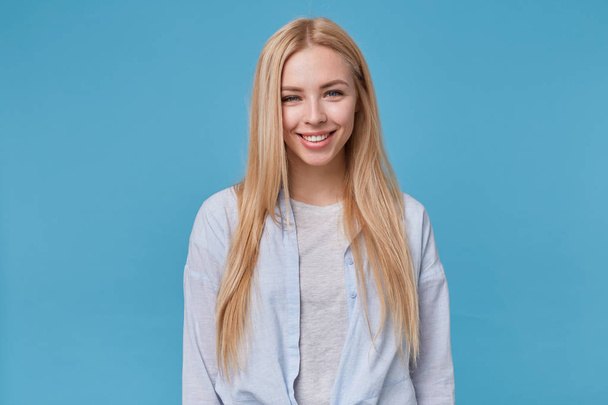 Porträt einer jungen blonden Frau mit lässiger Frisur, die vor blauem Hintergrund posiert, blaues Hemd und graues T-Shirt trägt und positive Emotionen zeigt, nachdem sie ein angenehmes Kompliment erhalten hat - Foto, Bild