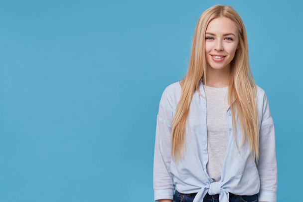 Beautiful young blonde female showing her pleasant emotions, keeping hands along her body, posing over blue background in blue shirt and grey t-shirt, looking to camera with charming smile - Foto, Bild