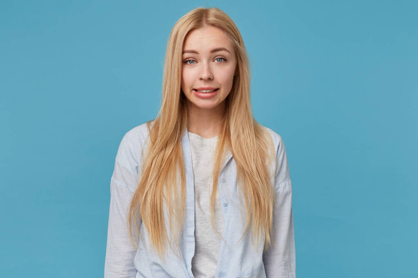 Portrait of blonde young attractive lady with casual hairstyle wearing blue shirt and grey t-shirt, looking at camera with confused face, posing over blue background with hands down - Fotografie, Obrázek