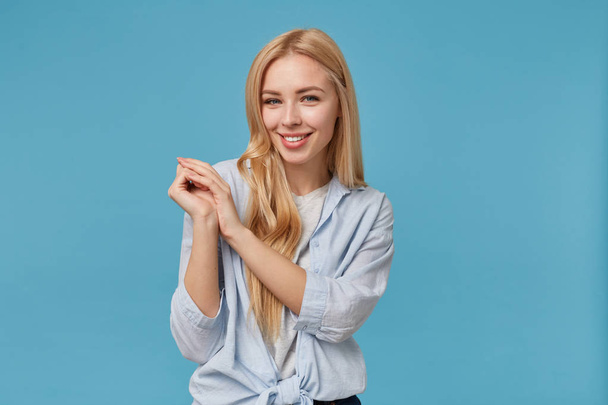 Okouzlující mladá blondýna s dlouhými vlasy žena zvedá ruce složené a usmívá se fotoaparát vesele, stojí nad modrým pozadím v ležérní oblečení, demonstrující své bílé dokonalé zuby - Fotografie, Obrázek