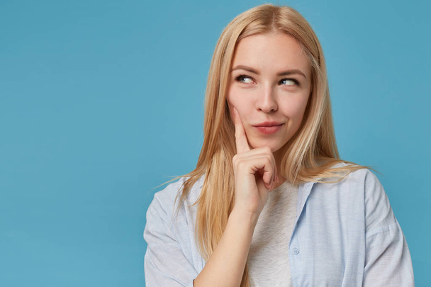 Close-up of beautiful young blonde lady with casual hairstyle looking aside thoughtfully, keeping forefinger on her cheek and smiling cunningly, isolated over blue background - Foto, Bild