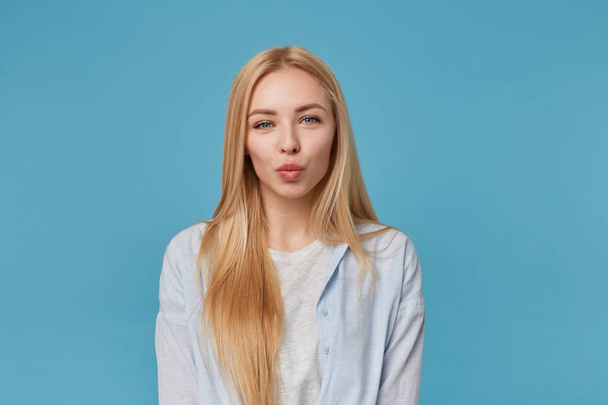 Indoor shot of beautiful blonde lady with casual hairstyle wearing blue shirt and grey t-shirt, posing over blue background with hands down, looking at camera positively with pursed lips - Photo, Image