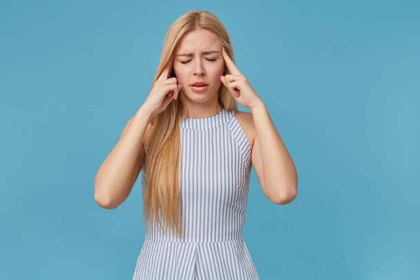Tired young blonde woman with long hair keeping forefingers on temples, closing eyes because of headache, posing over blue background in striped summer dress - Photo, Image