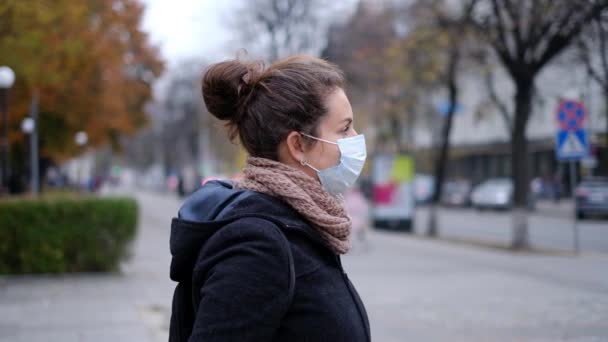 Μια νεαρή γυναίκα με ιατρική μάσκα στέκεται σε ένα δρόμο σε μια πόλη το φθινόπωρο.. - Πλάνα, βίντεο
