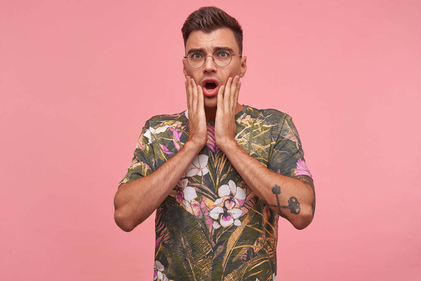 Красивый испуганный мужчина с закрытым ртом и руками, видящий что-то ужасное, одетый в очки и цветущую футболку, изолированный на розовом фоне
 - Фото, изображение