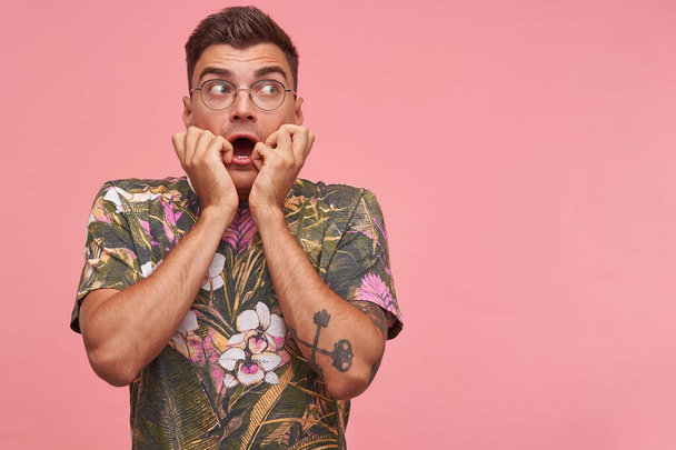 Portrait of scared young short haired male with hands on his face, being afraid of something, looking frightened aside, posing over pink background - Photo, Image