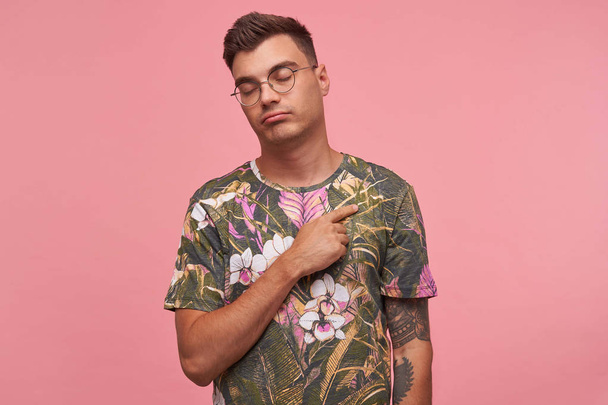 Charming young man with trendy haircut standing over pink background with eyes closed, keeping hand on his chest, being calm and balanced - Foto, imagen