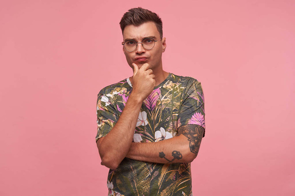 Portrait d'un jeune homme élégant et beau en t-shirt fleuri avec un visage sérieux, tenant son menton à la main et fronçant les sourcils à la caméra sur fond rose
 - Photo, image