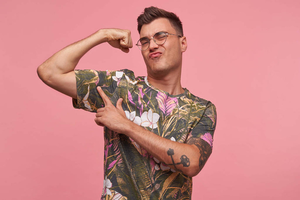 Estudio de tiro de orgulloso guapo tatuado joven en color casual camiseta señalando y mostrando su fuerte bíceps, aislado sobre fondo rosa
 - Foto, imagen