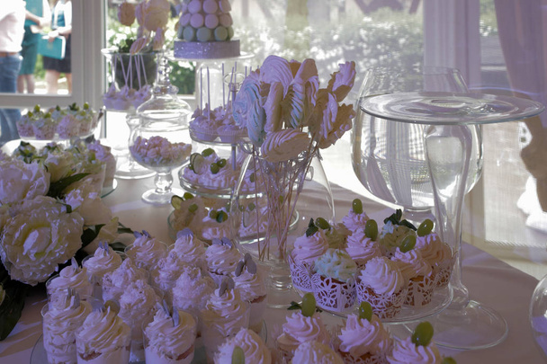 Barra de caramelo: mesa con un mantel blanco, cristalería, pasteles, dulces, macarrones, malvaviscos, crema, uvas
 - Foto, Imagen