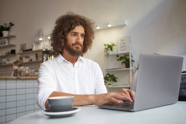 Varón severo que usa camisa blanca revisando correos electrónicos en línea, usando un portátil moderno, trabajando remotamente en el proyecto, manteniendo las manos en el teclado, tomando una taza de café
 - Foto, imagen