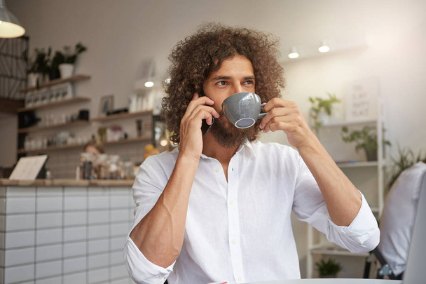 Close-up portrait of charming young male posing over cafe interior, drinking coffee while talking on phone, looking out window with calm face - Fotó, kép
