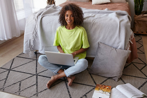 Wewnętrzny portret uroczej wesołej kobiety z brązowymi kręconymi włosami siedzącej na dywanie z geometrycznym nadrukiem, trzymającej laptopa na nogach i uśmiechniętej do kamery - Zdjęcie, obraz