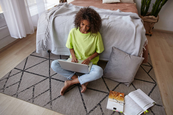 Молода приваблива темношкіра жінка з коричневим кучерявим волоссям, спираючись на ліжко в спальні, робить домашнє завдання з підручниками та сучасним ноутбуком
 - Фото, зображення