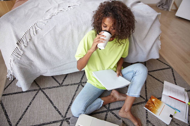 Jonge donkere vrouw met bruin krullend haar die thuis studeert, op tapijt zit met geometrische print, koffie drinkt en naar haar notities kijkt - Foto, afbeelding