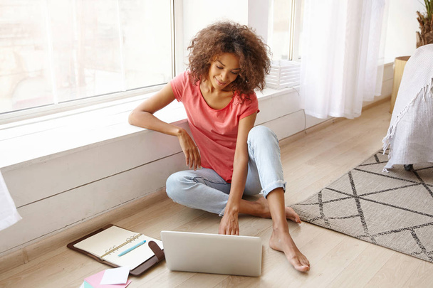 Indoor shot of attractive dark skinned woman leaning on windowsill while sitting on floor, working remotely from home with modern laptop and notebook, positive emotions - Фото, зображення