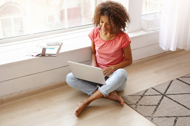 Jonge vrij donker gevilde krullende vrouw werkt op afstand van huis, zit op de vloer in de buurt van raam met laptop op benen, kijkt naar het scherm en glimlacht iets - Foto, afbeelding
