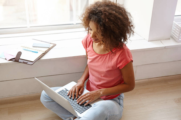 Retrato de una joven bonita mujer rizada con piel oscura sentada en el suelo con el ordenador portátil, manteniendo las manos en el teclado, posando sobre una amplia ventana, usando jeans y camiseta rosa
 - Foto, imagen