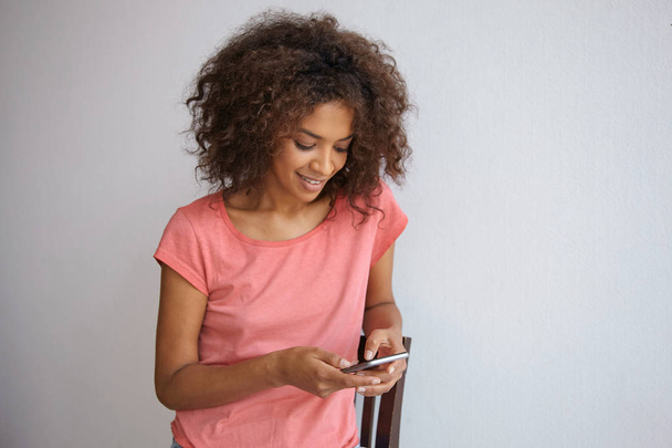 Közelkép gyönyörű fiatal sötét hajú göndör női gazdaság telefon a kezében és sms, nézi a képernyőn, és mosolygós lágy, visel alkalmi ruhák - Fotó, kép