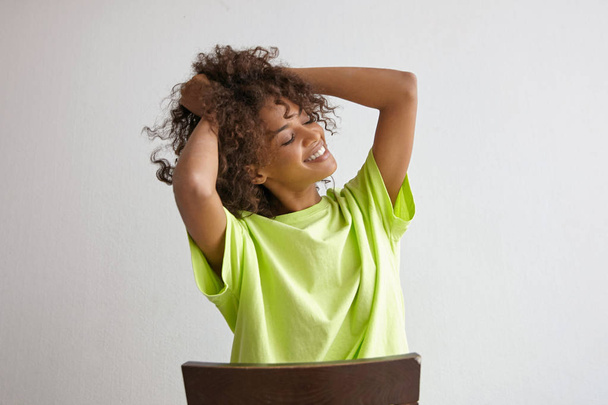 Indoor portrait of happy attractive curly female with dark skin sitting on chair over white background, holding her hair with hands, smiling cheerfully with closed eyes - Photo, Image