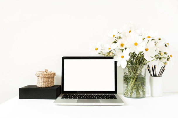 Business, freelance, blogger concept. Mock up, empty, copy space. Laptop standing on white table against white wall with daisies in vase, black box, straw basket.  - Photo, Image