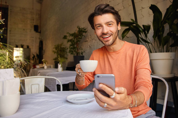 Attractive young dark haired man in sweater posing over cafe interior, holding srartphone in hand and looking to camera cheerfully, having cup of coffee - Zdjęcie, obraz