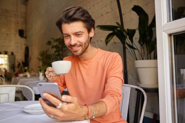 Indoor shot of handsome young man in peach-colored sweater drinking coffee in city cafe near window, holding phone in hand looking at screen - Valokuva, kuva