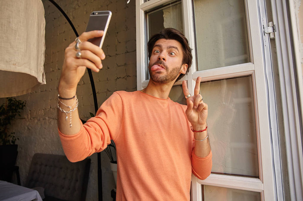 Funny indoor portrait of pretty bearded male holding phone in hand and leaning on window, making ridiculous faces and showing peace gesture, taking selfie with mobile phone - Photo, image