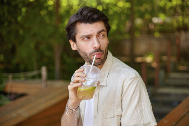 Außenporträt eines charmanten jungen bärtigen Mannes mit dunklen Haaren, der zur Seite schaut und Zitronen mit Stroh trinkt und über dem grünen Stadtgarten posiert - Foto, Bild