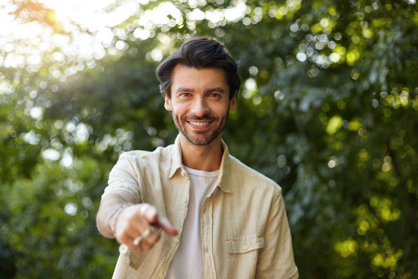 Портрет веселого молодого темноволосого мужчины, смотрящего в камеру с искренней улыбкой и поднявшего указательный палец, одетого в повседневную одежду, позирующего над зеленым парком
 - Фото, изображение