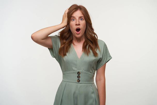 Indoor shot of surprised young woman with curly red hair, looking to camera with wide mouth opened, keeping palm on her head, isolated over white background - Fotografie, Obrázek