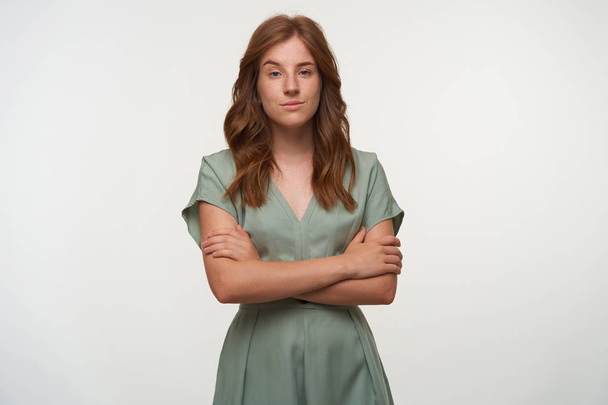 Portrait of pretty redhead female in pastel dress looking to camera with mysteriously smile, standing over white background with crossed arms - Photo, Image