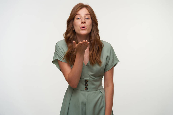 Studio photo of pretty young female with red hair wearing pastel dress, raising palm to her face and blowing air kiss to camera, isolated over white background - Zdjęcie, obraz