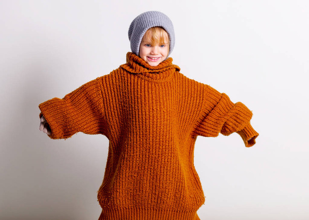 entzückendes kleines Mädchen, niedliches Kleinkind mit Strickmütze und großzügigem orangefarbenem Pullover, spielend und lustig lächelnd über die graue Wand des Studios. - Foto, Bild