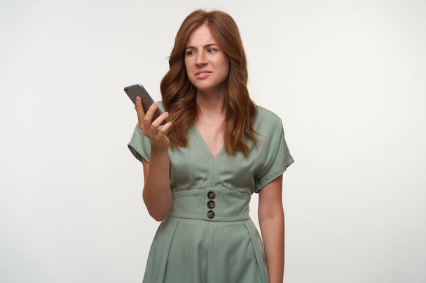 Спантеличена красива жінка з кучерявим рудим волоссям тримає смартфон в руці, дивлячись на екран зі збентеженим обличчям, позує на білому тлі
 - Фото, зображення