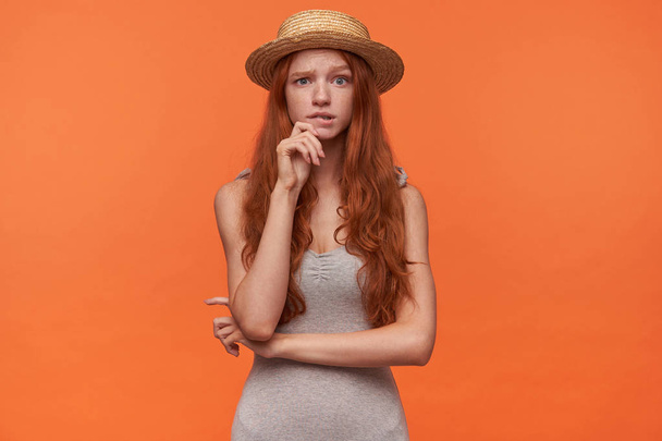 oszołomiona piękna młoda kobieta z falującymi lisimi włosami stojąca nad pomarańczowym tłem w szarym ubraniu słomiany kapelusz, trzymająca rękę na podbródku i niewątpliwie patrząca w kamerę - Zdjęcie, obraz