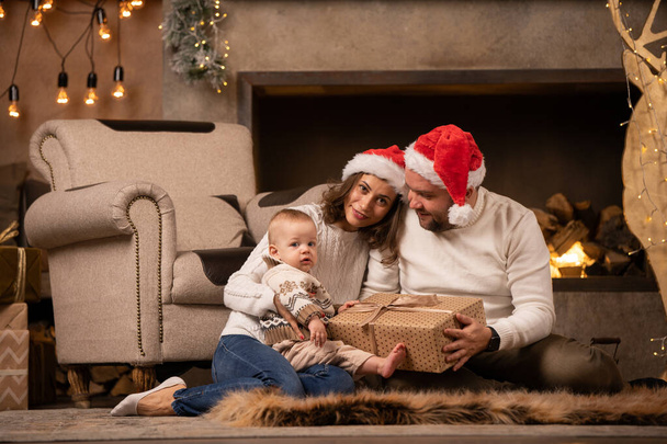 Picture of happy woman and man in Santas cap with sons at fireplace with garland in room with Christmas interior - Foto, Bild
