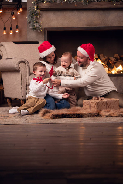 Image of parents in Santas cap with two sons sitting on floor by fireplace in room - Valokuva, kuva