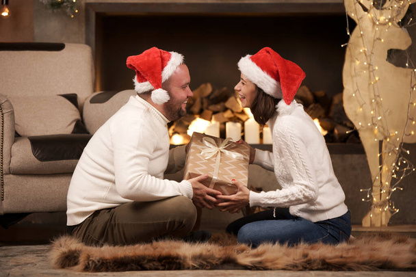 Picture from side of man and woman in Santas caps sitting on floor in room - Фото, изображение