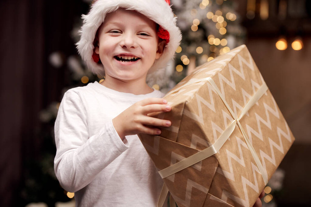Image of boy in Santas cap with gift in hands in room. - Foto, Imagen