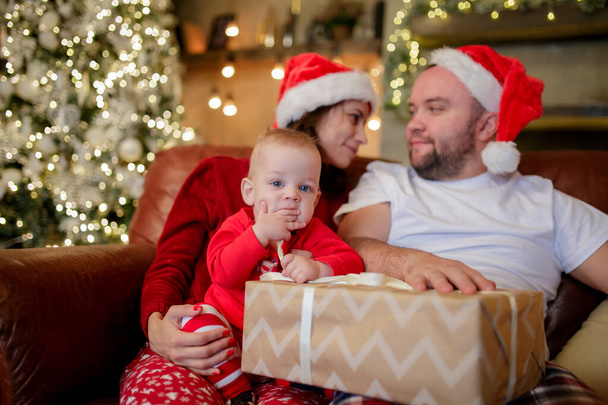 Obrázek mladých rodičů v čepicích Santas se synem sedícím na židli v pokoji . - Fotografie, Obrázek