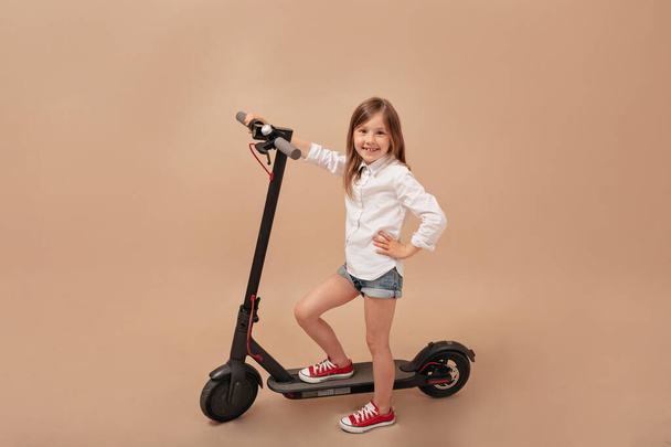Indoor frame of little cute girl posing at camera over isolated beige background with electric scooter - Foto, Bild