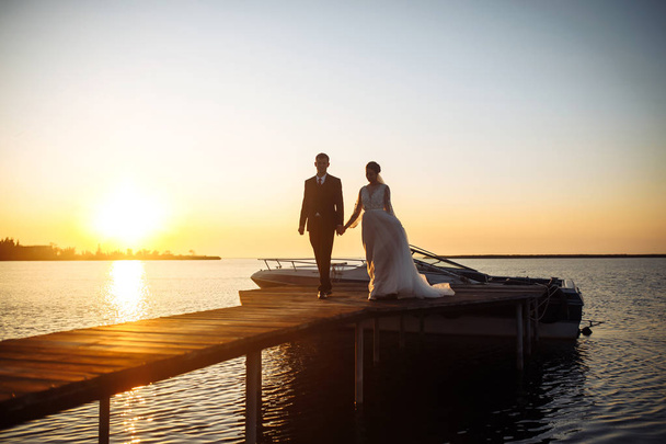 Beautiful bride and stylish groom together on the bridge against the background of the boat at sunset. Newlyweds tenderly hug, kiss and enjoy each other at sunset. Wedding. Love. Romantic moment.  - Foto, Imagen