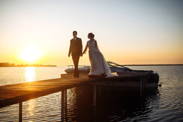 Beautiful bride and stylish groom together on the bridge against the background of the boat at sunset. Newlyweds tenderly hug, kiss and enjoy each other at sunset. Wedding. Love. Romantic moment.  - Фото, изображение