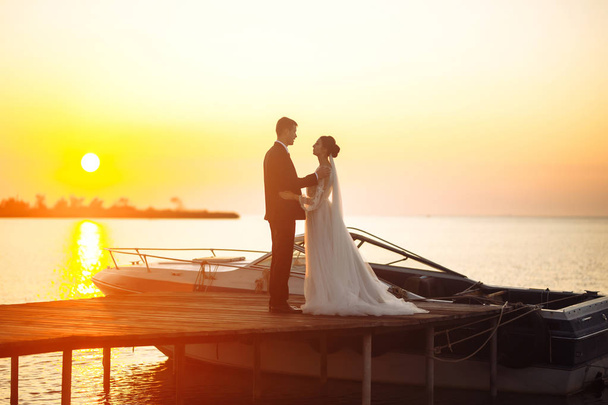 Beautiful bride and stylish groom together on the bridge against the background of the boat at sunset. Newlyweds tenderly hug, kiss and enjoy each other at sunset. Wedding. Love. Romantic moment.  - Foto, immagini