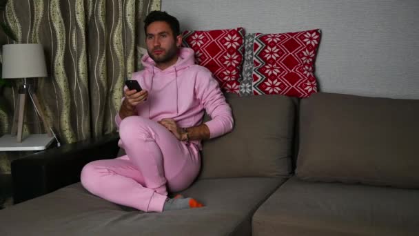 A young man in a gloomy mood and pink pajamas sits at home on the sofa, watches an uninteresting TV show and waves his hands with a remote control in displeasure. - Séquence, vidéo