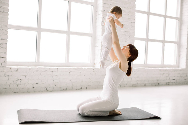 Sport, motherhood and active lifestyle concept - side view of young mother workout together with her baby sitting on the black mat in big light room. Mother having fun and playing with her little girl - Foto, afbeelding