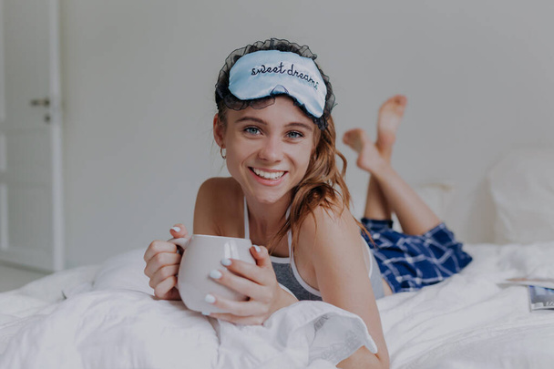 Chica feliz usando ropa de dormir y máscara de dormir acostado en la cama con una taza y sonriendo
  - Foto, imagen